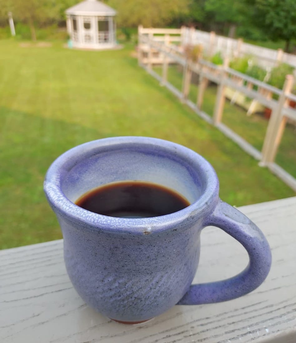 A close-up on a clay, purple mug of coffee from above. Blurred in the background is a big green yard with a garden and a gazebo in the distance.