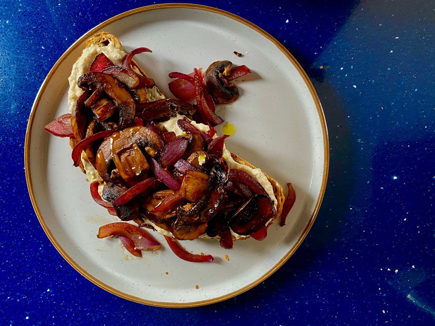 Piece of toast topped with a creamy beige coloured spread, with pieces of red onion and sliced mushrooms scattered over the top. The food is served on a round plate placed on a blue countertop