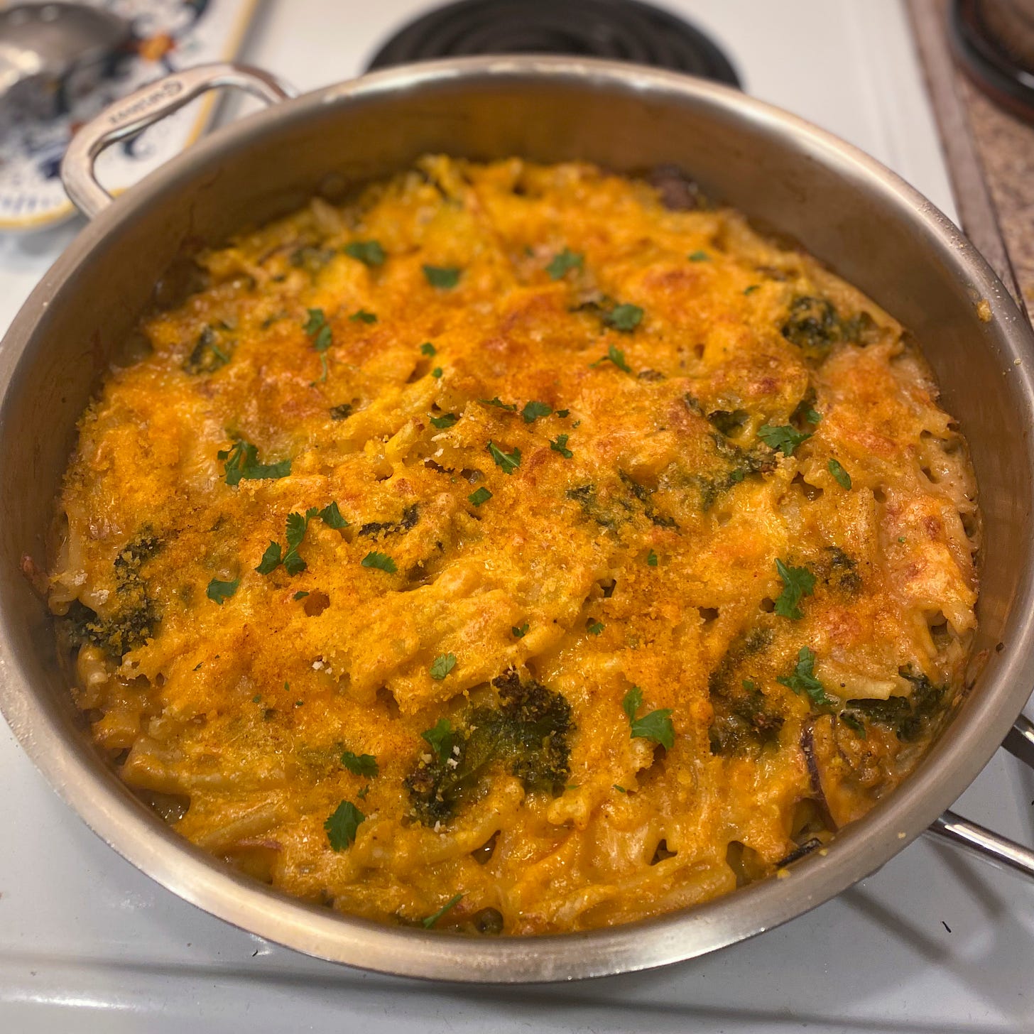 A large stainless steel pan full of yellow-orange mac & cheeze, with bits of kale throughout. On top is a browned crust of cheeze and breadcrumbs, and it is sprinkled with cilantro.