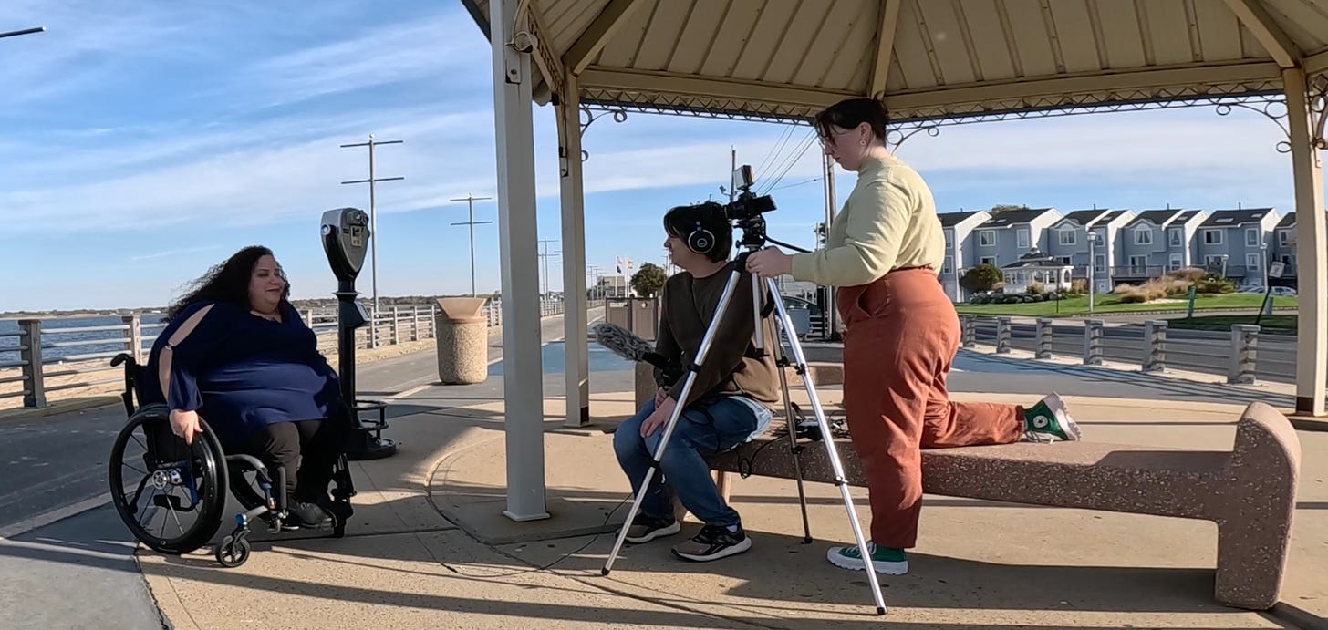 Along a beachfront promenade, a woman in a wheelchair faces a seated man holding a microphone and a woman behind a camera atop a tripod.