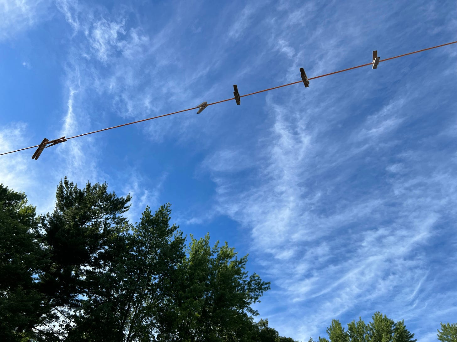A clothesline and clothespins against a mostly clear sky, a hew high clouds, and some evergreen trees