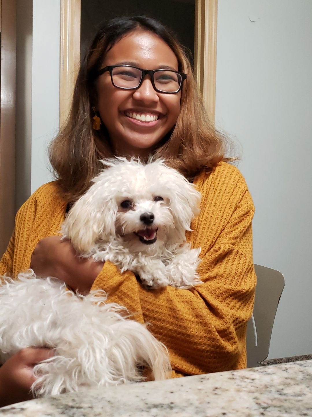 Melissa sits in a chair next to a table. She is holding a fluffy, small white dog and smiling widely. The dog is also smiling.