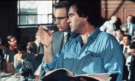 This image shows JFK's director and co-writer Olive Stone (right) directing actor Kevin Costner (playing the role of New Orleans District Attorney Jim Garrison, left) during the film's long courtroom sequence. Stone gestures with his right hand while holding either the movie's screenplay or storyboards in his left hand. Costner listens intently to Stone, while background performers dressed in 1960s' period costumes sit in the courtroom set's gallery behind Stone and Costner. This image shows JFK's director and co-writer Olive Stone (right) directing actor Kevin Costner (playing the role of New Orleans District Attorney Jim Garrison, left) during the film's long courtroom sequence. Stone gestures with his right hand while holding either the movie's screenplay or storyboards in his left hand. Costner listens intently to Stone, while background performers dressed in 1960s' period costumes sit in the courtroom set's gallery behind Stone and Costner.