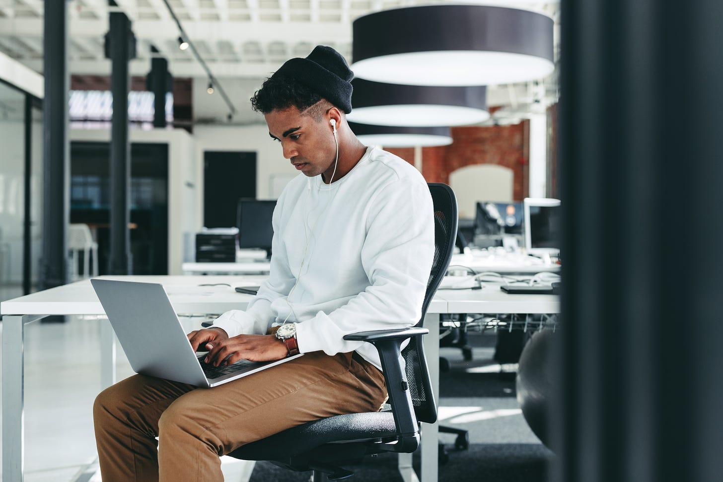 A Black, male tech worker wearing a black beanie, white sweatshirt, and brown pants sitting alone at a laptop in an expansive, modern office.