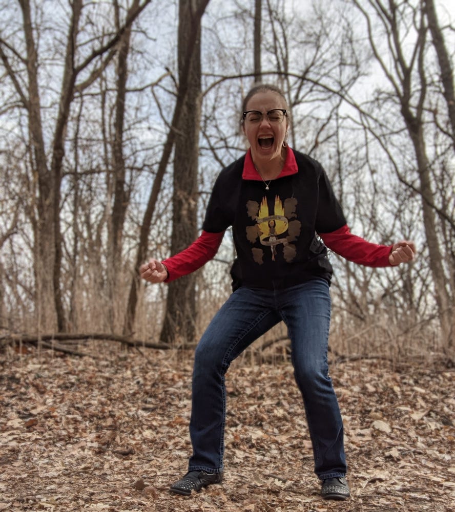 A person in a red and black shirt and jeans standing in a forest with trees behind them and leaves on the ground. The person is making fists with their hands and is screaming.
