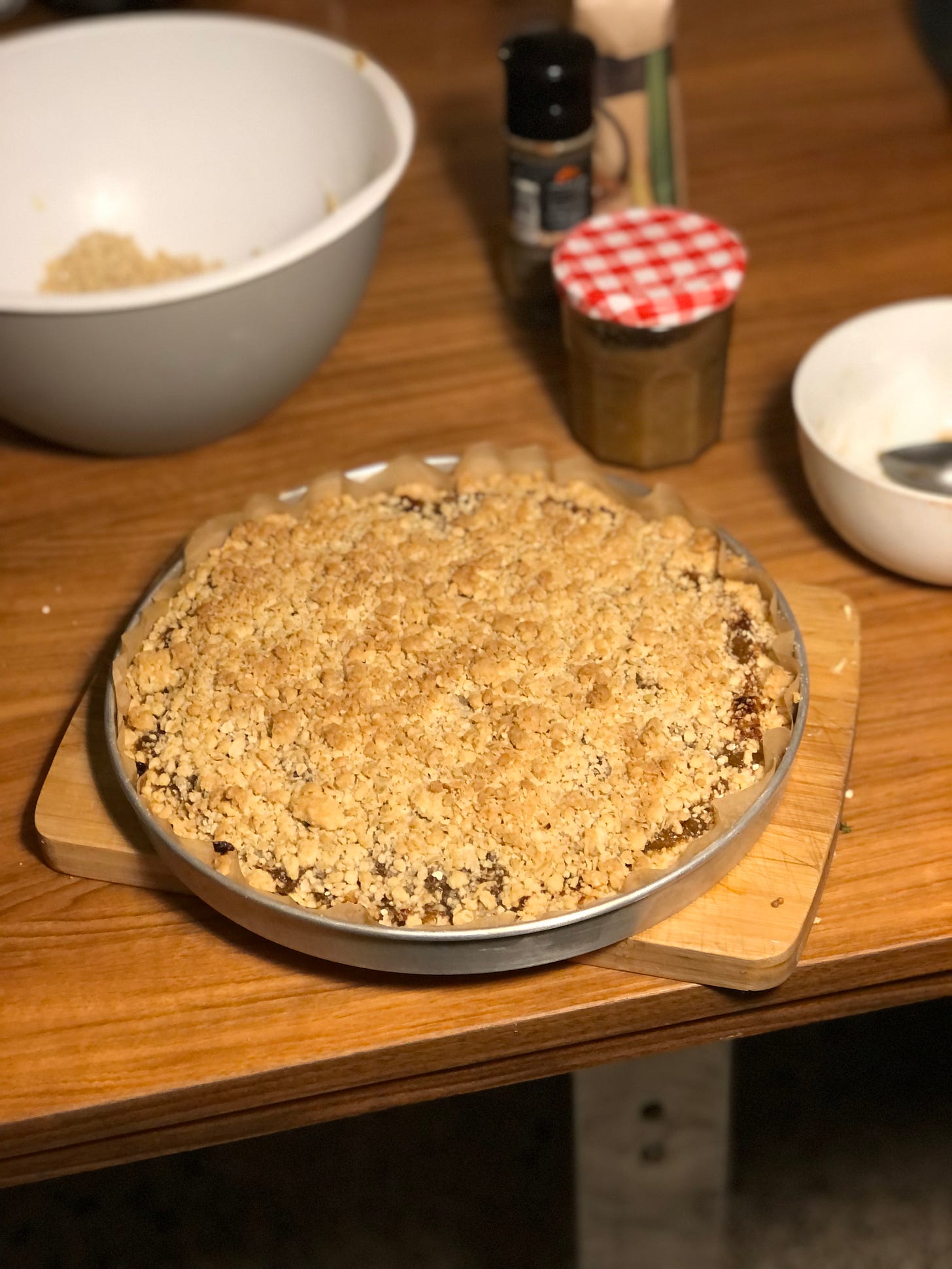 A photo of a crumble-topped dessert, blonde-brown on top, in a wide shallow round silver baking tray, on a breadboard on a table. Out of focus, in the background, a large mixing bowl and some spice jars. 