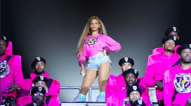 Beyonce at Coachella, standing in a pink sweatshirt and jean shorts surrounded by Black male drummers in pink uniforms. Beyonce at Coachella, standing in a pink sweatshirt and jean shorts surrounded by Black male drummers in pink uniforms.