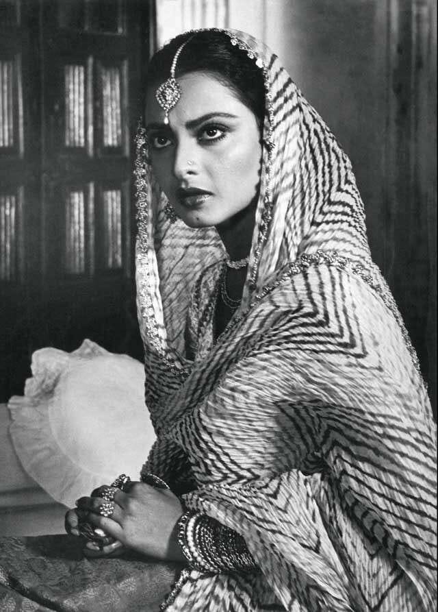 A black and white picture of actress Rekha in costume as the character Umrao Jaan, staring intensely towards something. She is wearing salwar kameez with a dupatta draped around her head, and jewellery around her neck, and on her ears and head.