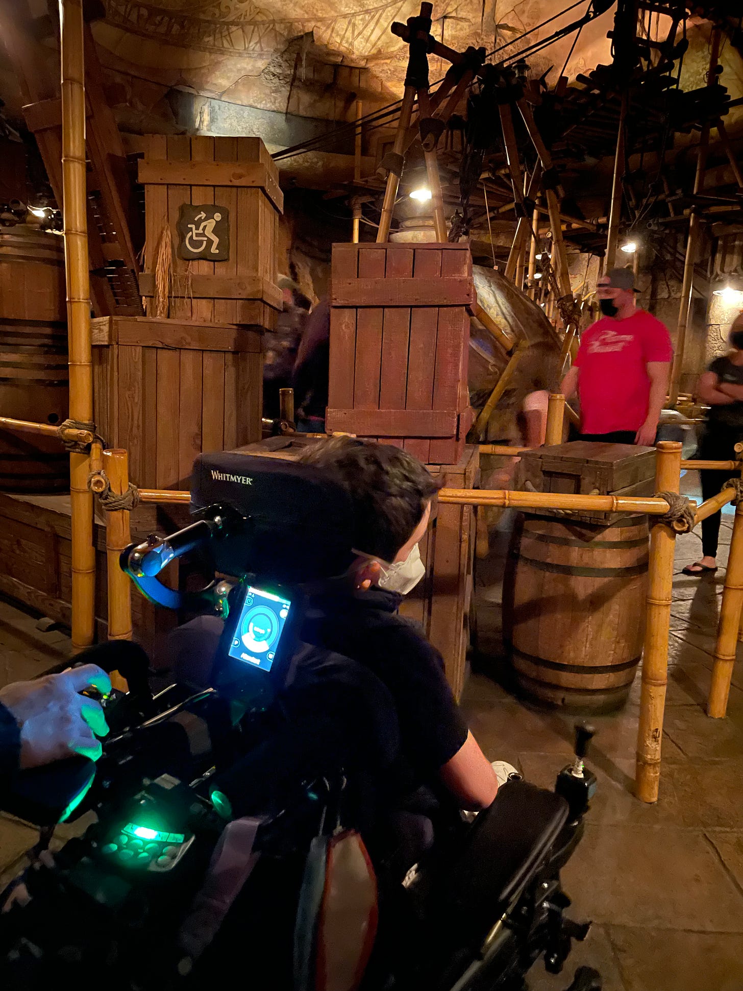 A wheelchair transfer sign on crates inside a cavernous hall made to look like the Indiana Jones movie, a boy in a powerchair waits at a rope line. 