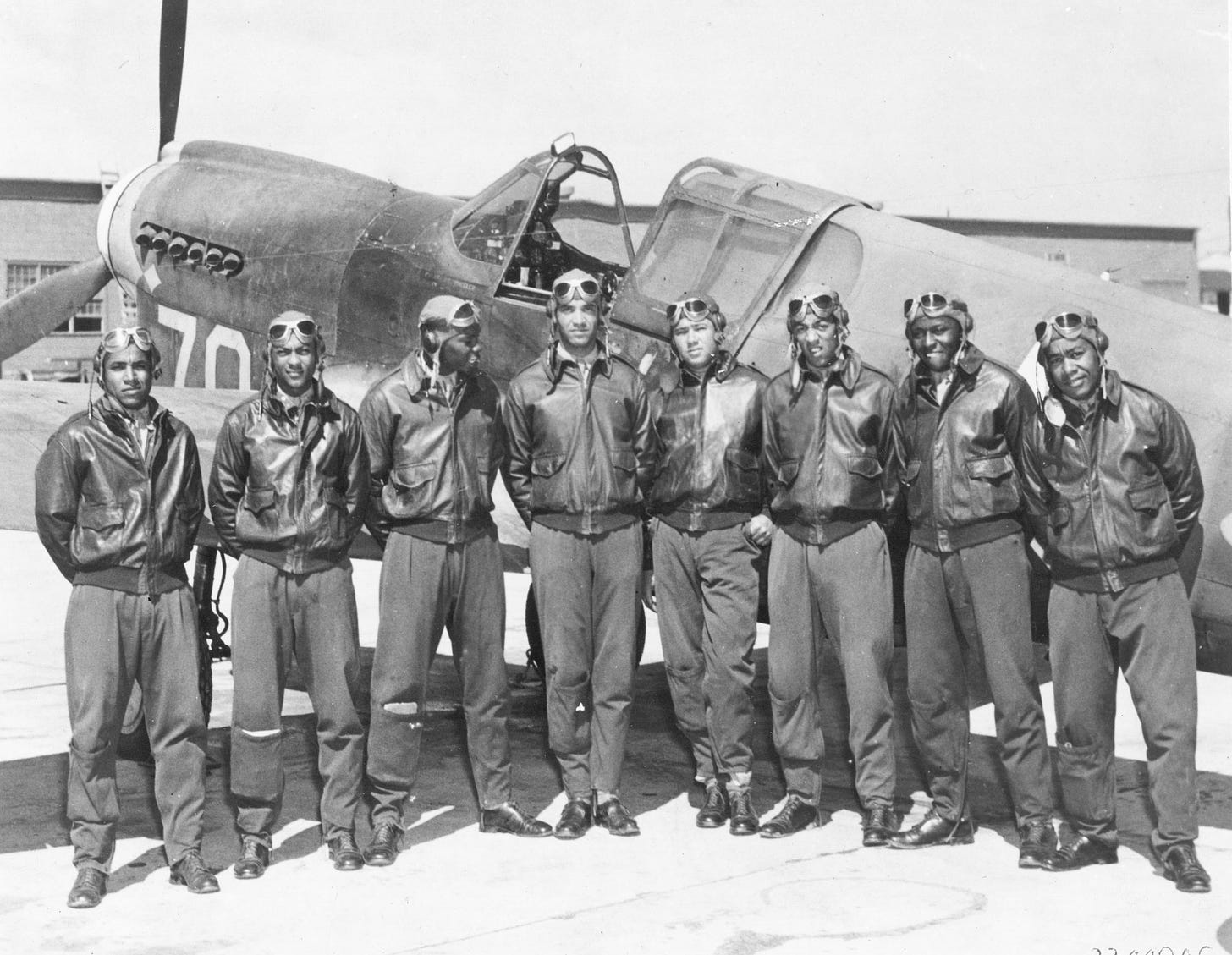Eight Tuskegee pilots are pictured, in flight suits, in front of a plane.