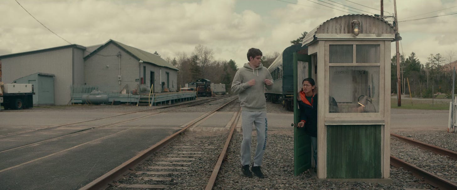 A still from the film 'The Half of It'. Ellie and Paul are at the train station where Ellie and her dad work. Paul is wearing a grey tracksuit. Ellie is wearing glasses, a navy jacket and blue jeans.