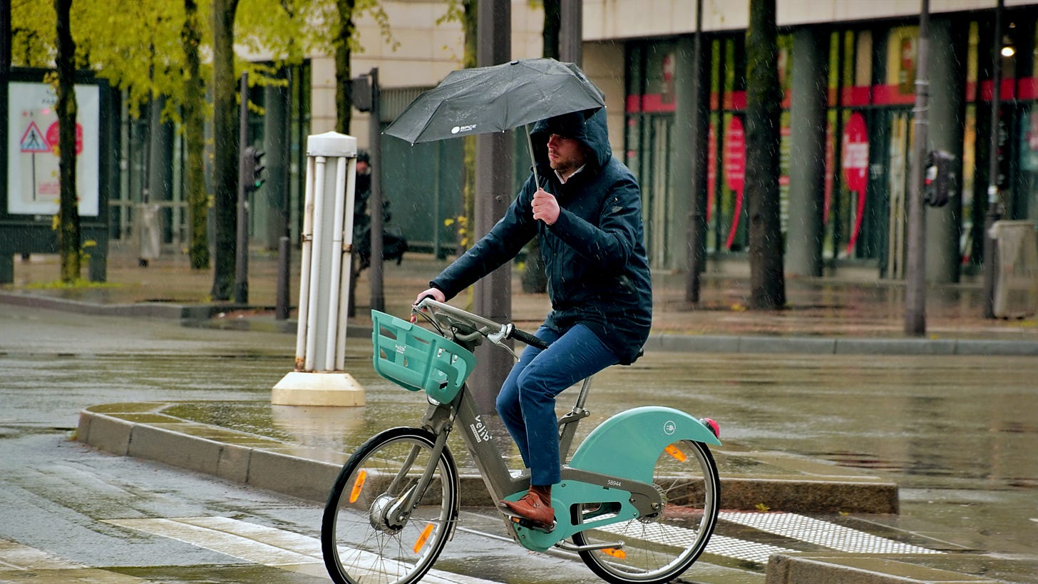 Man wearing hooded jacket holding umbrella on bicycle at crosswalk in the rain