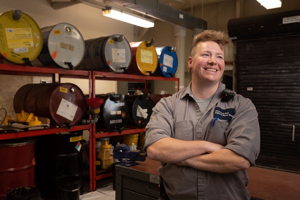 Portrait of Kate, smiling as she looks off camera, arms folded, standing in front of a maintenance shop rack of colorful 55-gallon drums.