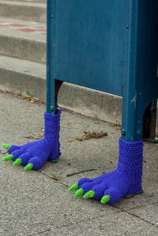 Close-up of a blue Post Office mail box with hand knitted blue booties with green talons made to look like furry monster's feet.