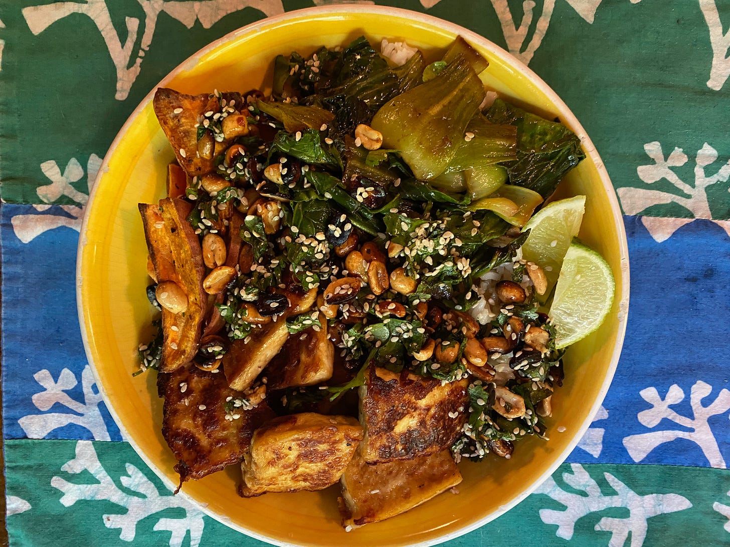 A wide yellow ceramic bowl holding sweet potato wedges, crispy brown tofu squares, sautéed bok choy, and lime wedges, with peanuts and sesame seeds scattered on top.