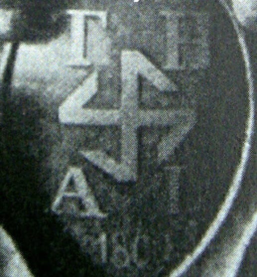 A black and white picture showing an oval design showing a hooked cross design. Above the hooked cross reads “T” and “H” and below reads “A” and “L”. Below that is the date: “1869 although that isn’t very clear.