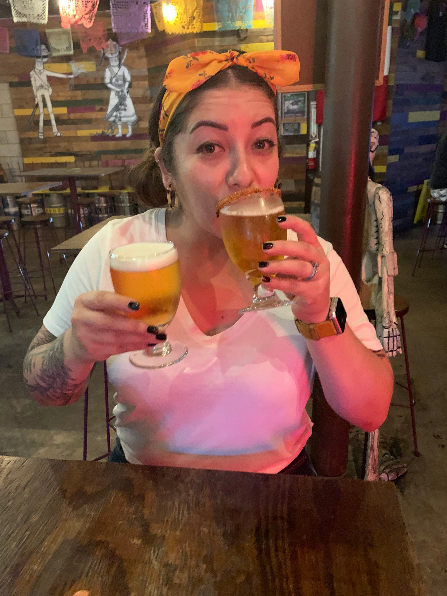 Chicana woman wearing an orange headband and holding a beer in each hand, taking a sip from each one.