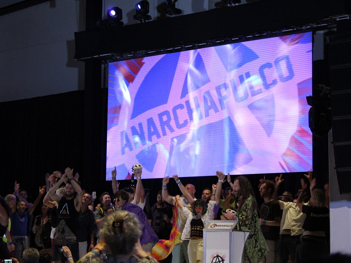Crowd of attendees at Anarchapulco with their hands in the air. Anarchy symbol and text Anarchapulco displayed on giant...