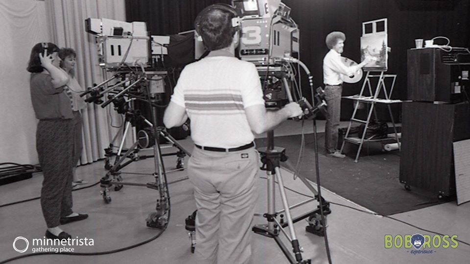 Wide view of Bob Ross' painting and filming studio with Bob at his easel and three crew members filming
