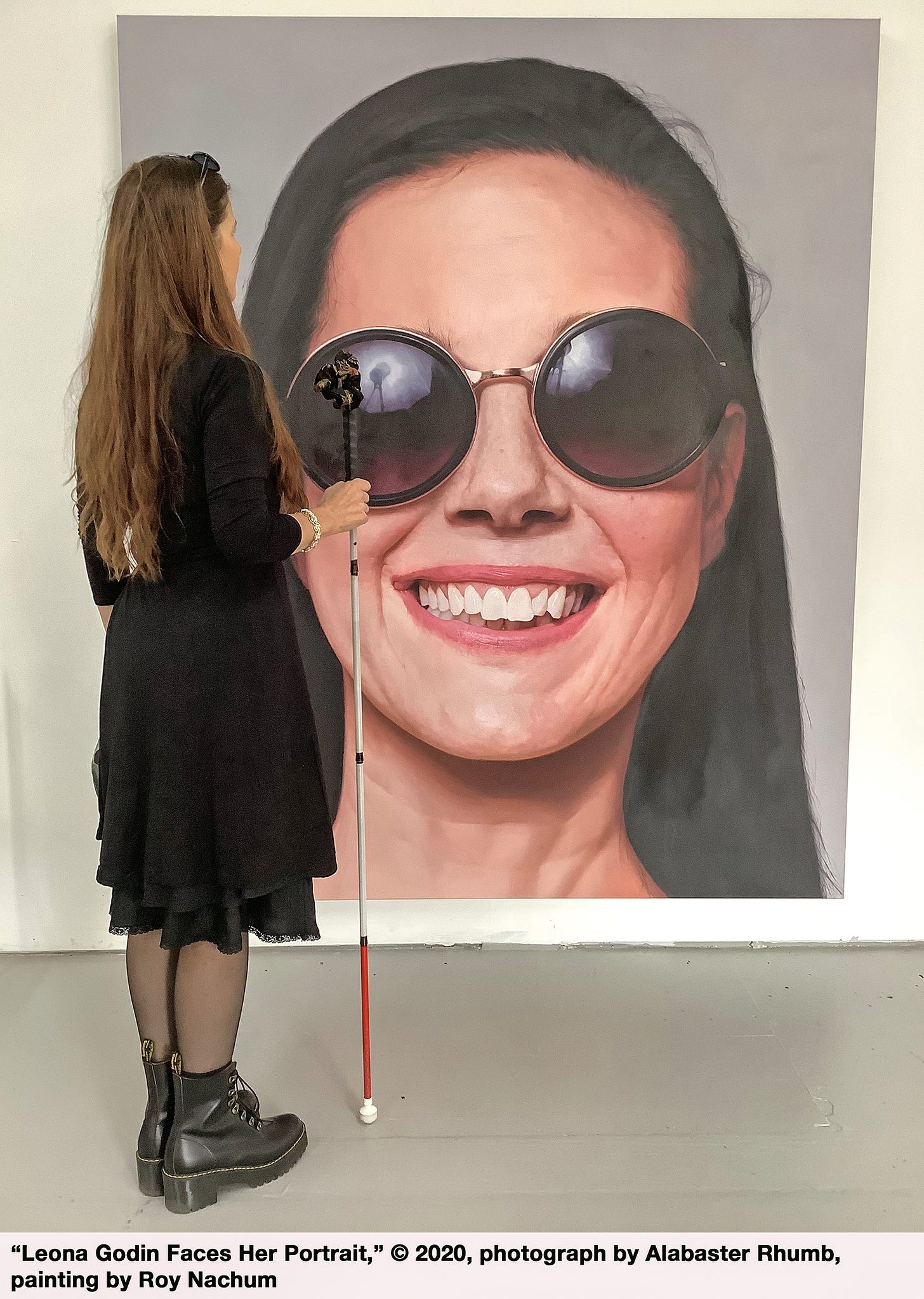 By M. Leona Godin: I’m standing to the side of and facing a six-foot canvas on which is painted a hyper-realistic close-up of my head. In the painting I’m wearing purple and gold sunglasses (which sit slightly askew, and which reflect the lighting umbrellas) and smiling. In the studio I’m standing in a black wrap dress, wearing black Dr Martens, and holding Moses, who bisects the painting from the sunglasses downward. My black sunglasses are perched on my head. Author image credits - The painting is by Roy Nachum, and the photograph was taken in Roy’s studio by Alabaster Rhumb.