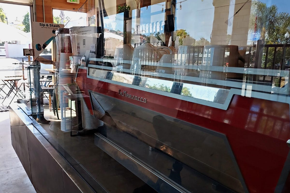 A close-up of a red and silver La Marzocco espresso machine through safety plexiglass. White coffee cups are stacked on top and it leads the eye out towards the open garage door and a view of the patio tables on the sidewalk.