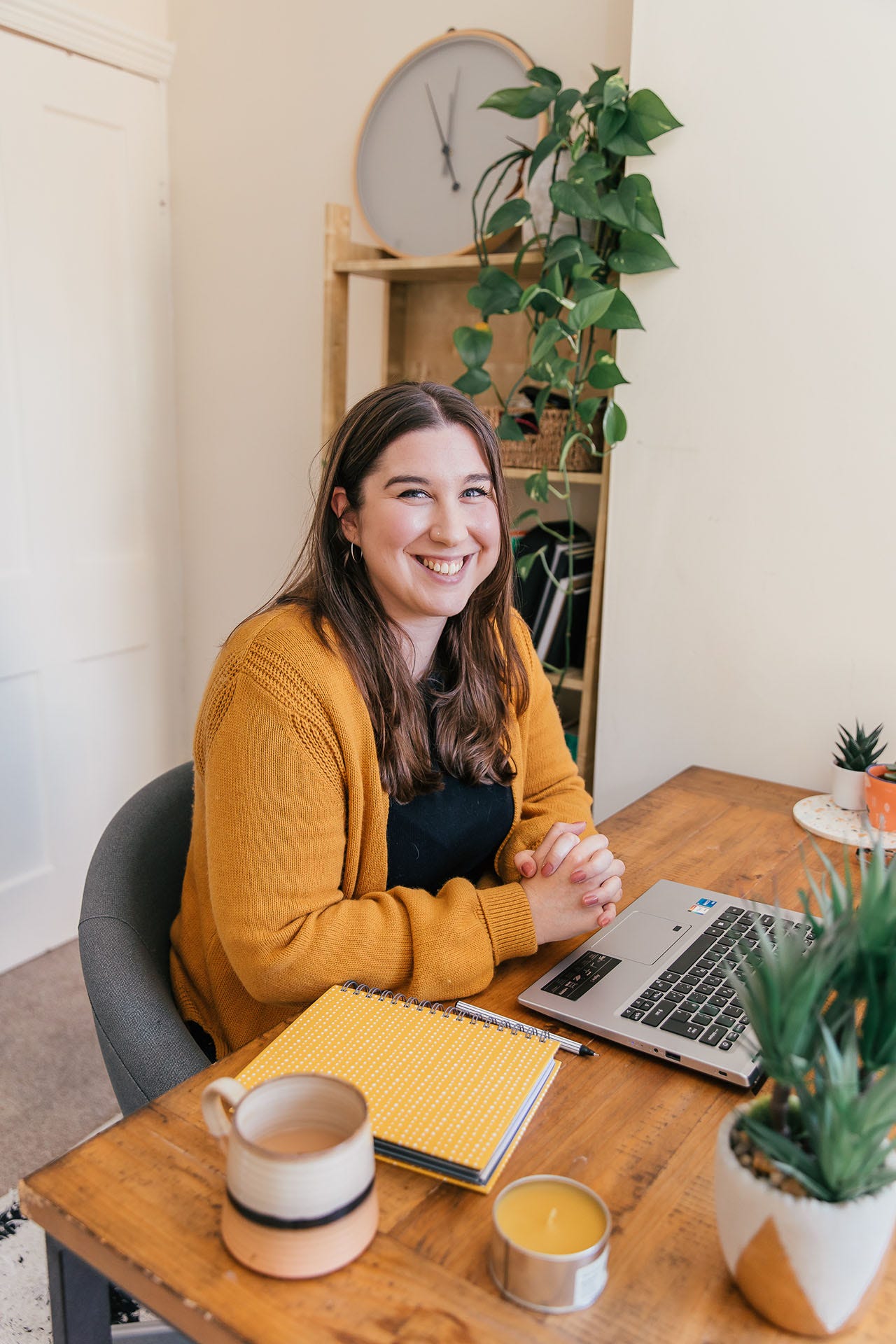 Rachel Baker, AKA The Ethical Copywriter, sits at a wooden desk. There is a laptop in front of her, and a yellow notebook, cup of coffee, yellow candle and plant to her right. There are several smaller plants to her left. She is looking at the camera and smiling. 