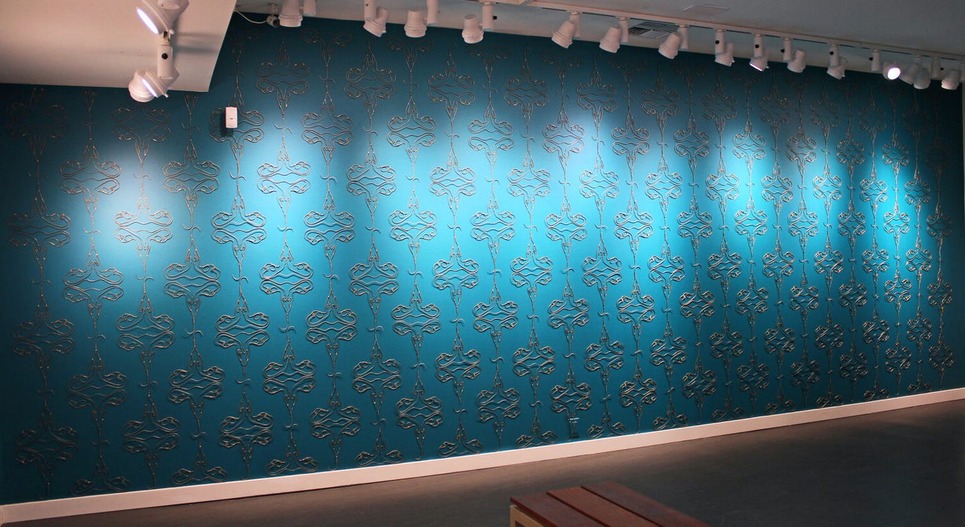 a long wall painted dark teal, lit by track lighting. Installed on the wall, to look like the pattern of wall paper, are a series of identical, repeating hand-formed wire sculptures. Each one looks like someone took a wire hanger and made it into a diagram of the female reproductive system.