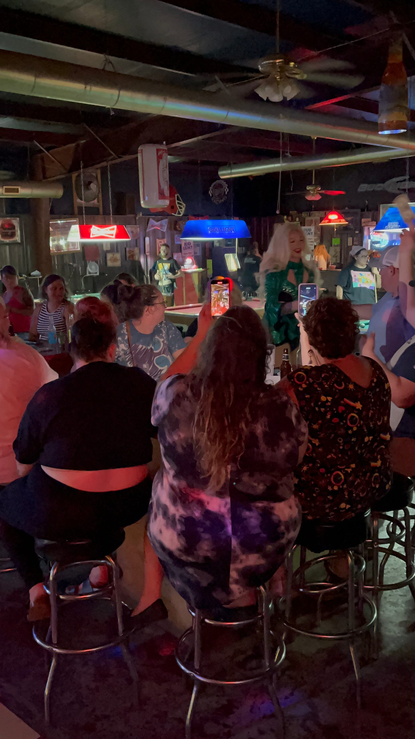 Multiple people sit on bar stools in a dark bar room filled with neon lights as a drag queen performs and is being photographed with smart phones.