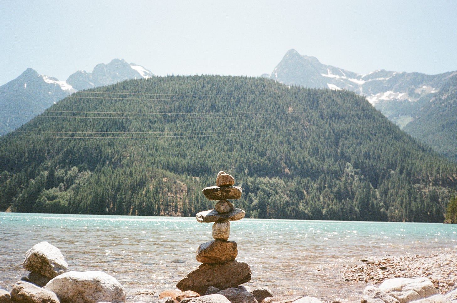 A cairn in the foreground of Diablo Lake wa, with a hill behind the lake and the Cascade Mountains behind it. 