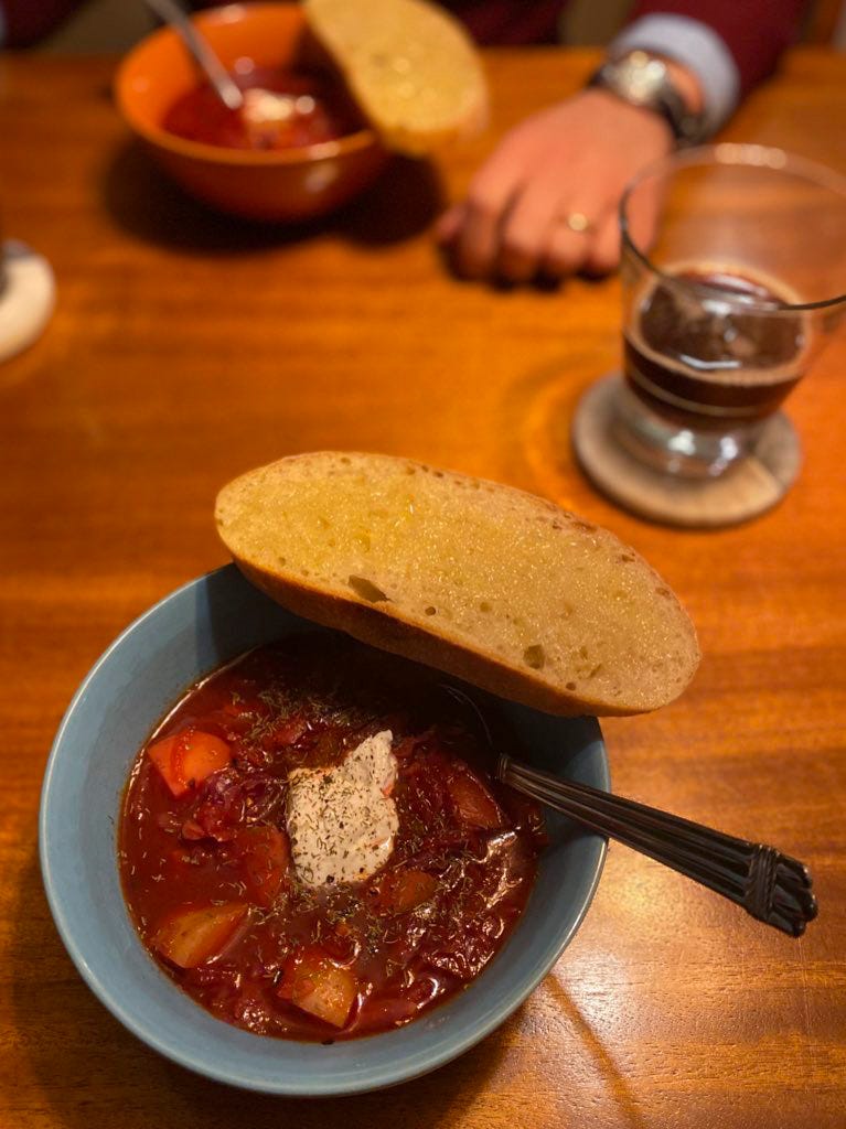 A blue bowl of dark pink borscht with sour cream and dill on top. A slice of buttered sourdough rests on the rim of the bowl, and there is a coaster to the right with a half-full glass of dark beer. In the background is an orange bowl featuring the same. Jeff's hand rests on the table beside it.