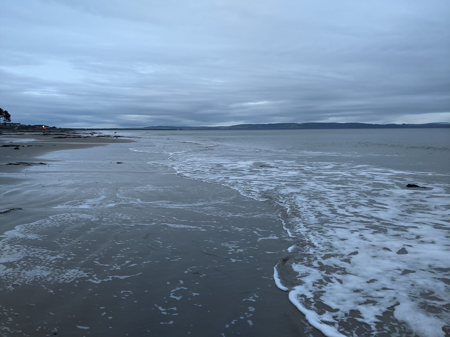 Sandy beach with frothy flat waves approaching from the right.  Grey sky above.