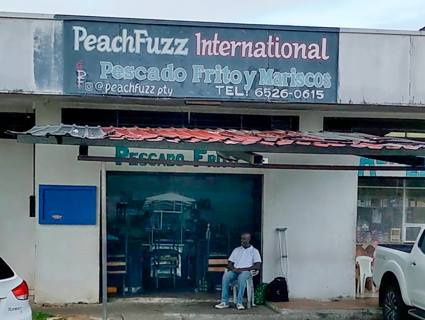 A man wearing a white polo shirt and blue jeans sits under the awning of a restaurant. The restaurant sign reads, PeachFuzz International, Pescado Frito y Mariscos