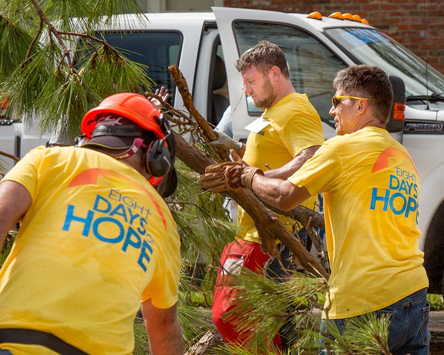 Three men in yellow shirts and safety equipment clean up fallen tree branches outside a home.