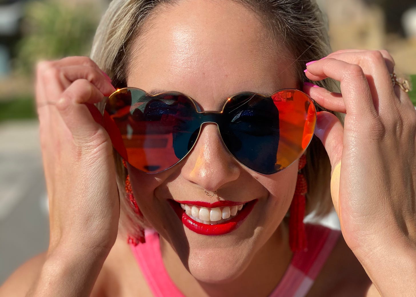 A woman with red lipstick smiles widely as she touches her red, heart-shaped sunglasses