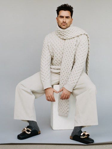 Photo of Dan Levy in a cream-color knit sweater, matching scarf, and cream pants, and Birkenstock-like slides.