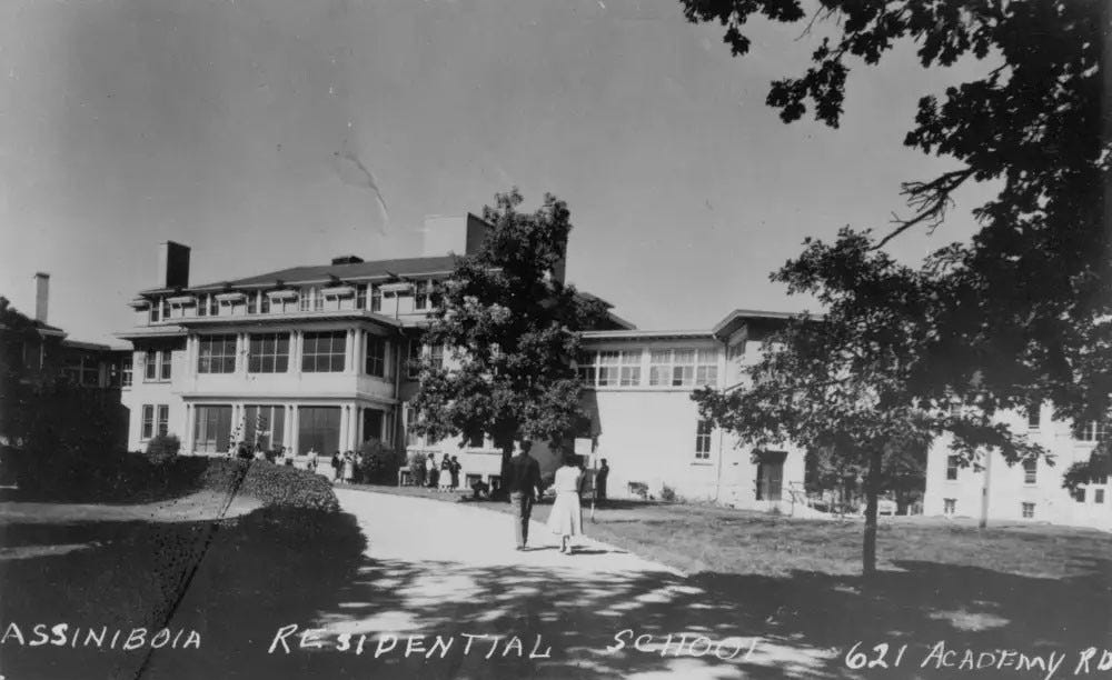 Black and white picture of three-story Assiniboia Residential School surrounded by trees, a road, a hedge, and a manicured lawn. Two people are walking towards the school, holding hands.