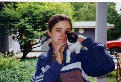 A young person with long, dark brown hair pulled back into an awkward ponytail, has a small kitten on their shoulder. They are dressed in a vintage Adidas track jacket.