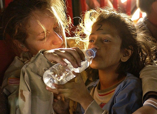 A white women in a khaki uniform holds a plastic bottle of water to a little girls mouth while hugging the child around her shoulders. The child’s face is tear-stained. She is light-skinned and light-haired enough to seem like a white girl. The girl is drinking from the water bottle. Both people are back lit by the sun making their hair seem golden. Another person is in the background on the top right hand corner but they are blurred with only the back of their neck being visible.