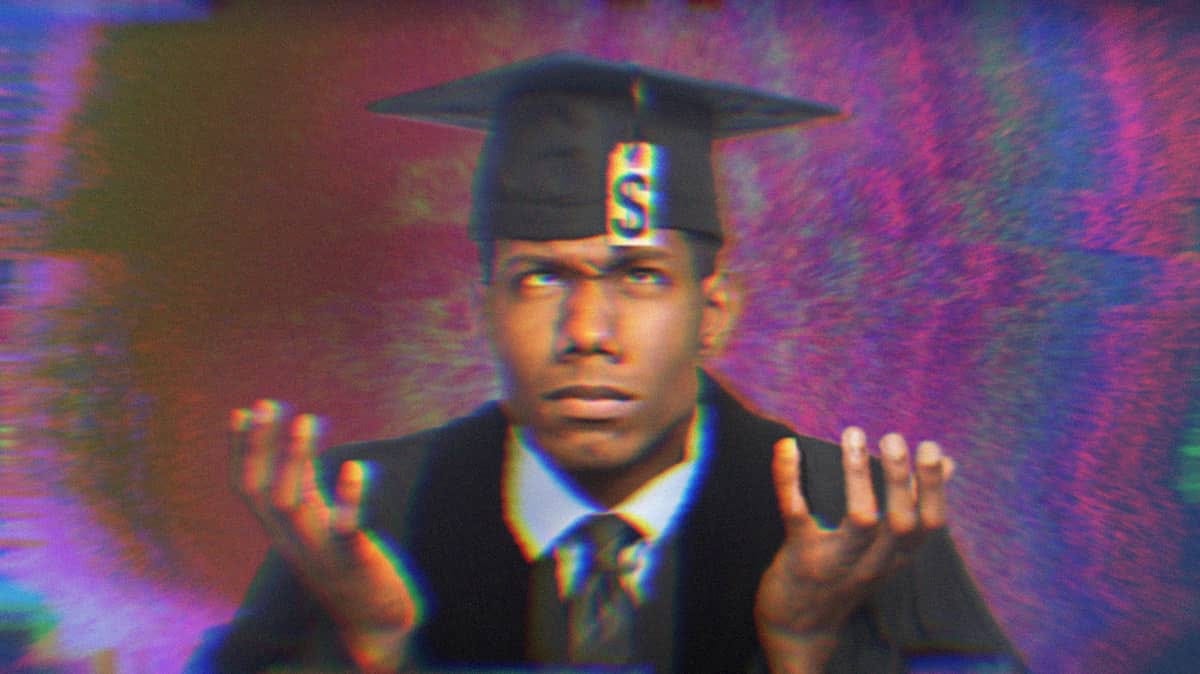 A young graduate wearing a cap and gown, shrugging and looking up at his graduation tassle which is a tag with a dollar sign on it.