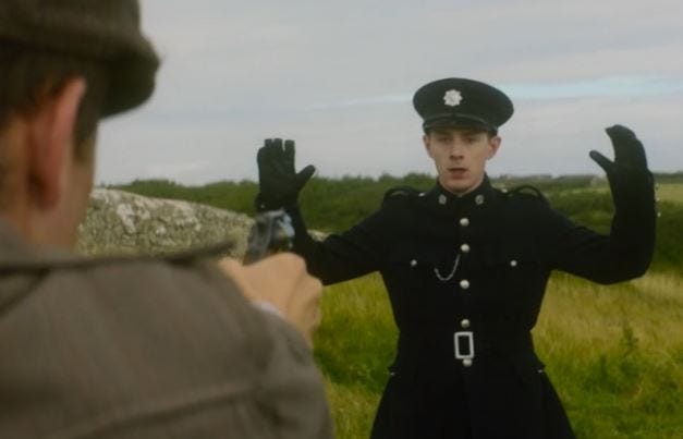 This image shows the character known only as Brendan (played by Evan McCabe), a twentieth-century Irish policeman, holding his hands up as a thief points a pistol at him. The thief is turned away from camera, so we only see his back and the pistol in his right hand. This image shows the character known only as Brendan (played by Evan McCabe), a twentieth-century Irish policeman, holding his hands up as a thief points a pistol at him. The thief is turned away from camera, so we only see his back and the pistol in his right hand.