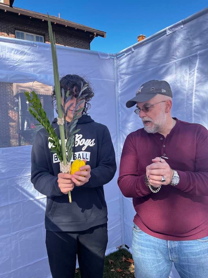 A white teenage boy holds a lulav and an etrog in a white-walled sukkah while a middle aged white man shows him how to shake them.