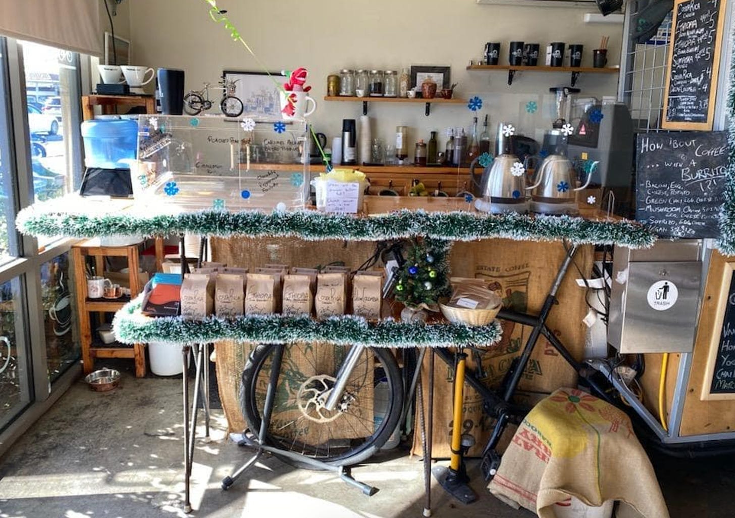 The inside of Coffee Cycle. The coffee cart is covered with green frosted holiday tinsel with bags of fresh coffee beans on a shelf below the counter.
