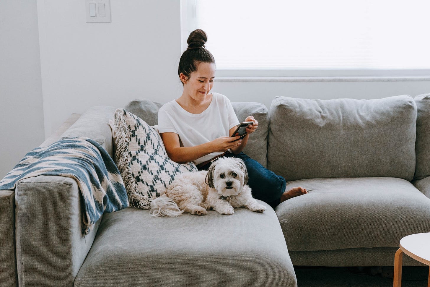 A woman smiles while looking at her phone and sitting next to her dog on the couch.