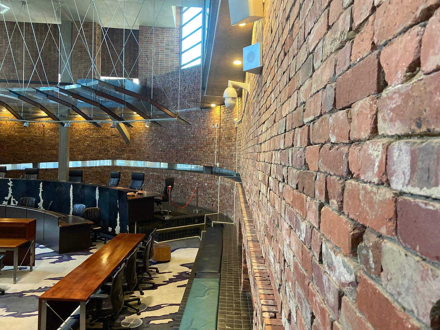 South African Constitutional Court chambers. Walls constructed with bricks from old prison. Eye-level window that shows only the feet of people walking by on the sidewalk. Author photograph. July 2, 2022.