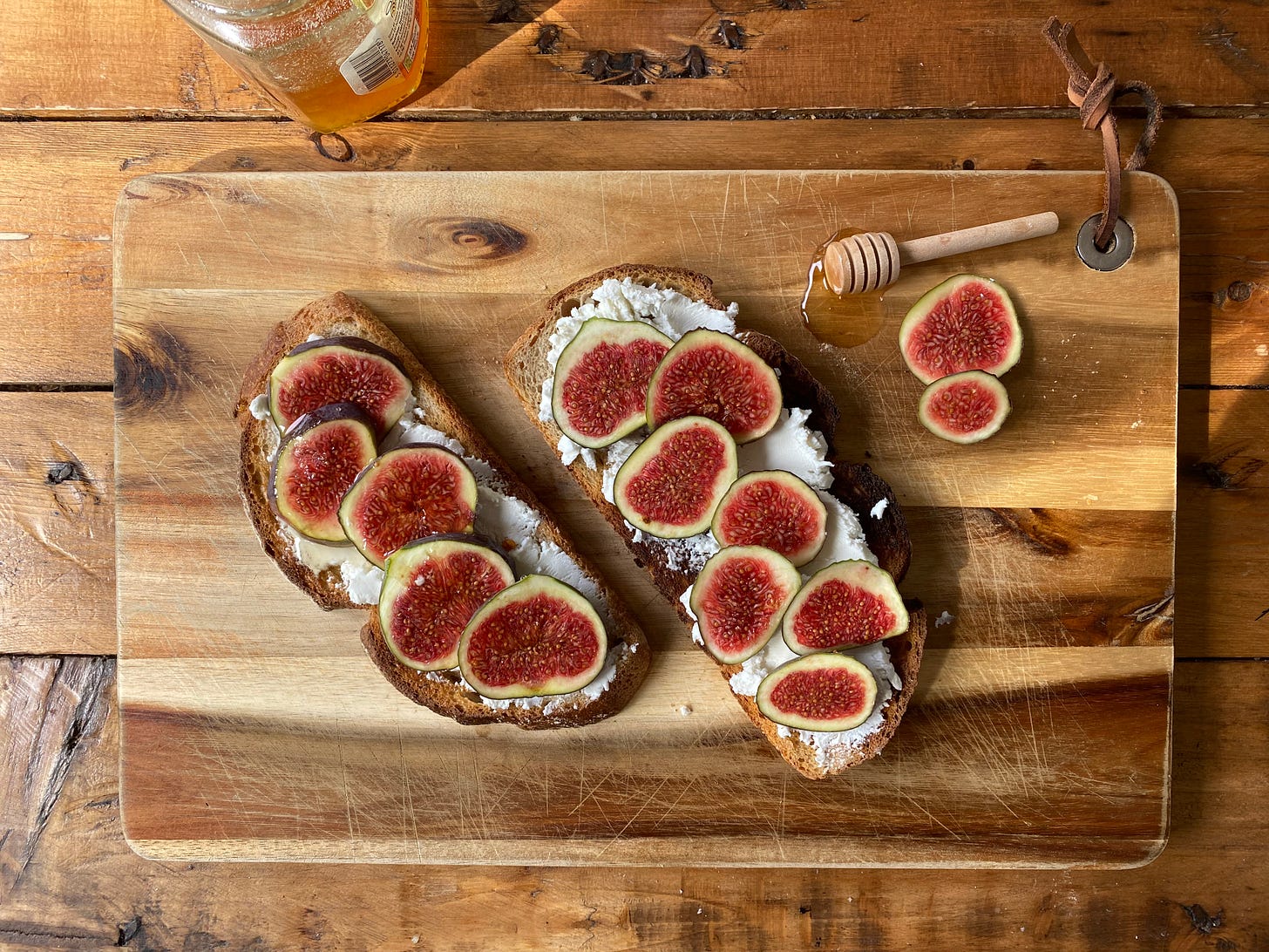 Wooden board topped with two pieces of bread. White cream cheese and and sliced figs are on the bread. A pot of honey to the side. 