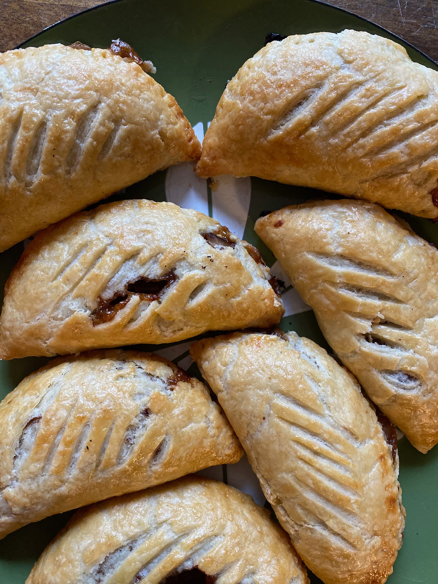 A close up of seven apple-onion turnovers on a green plate. They are half circles, with slashes running across their golden-brown tops. A few of them have burst open, showing the filling inside.