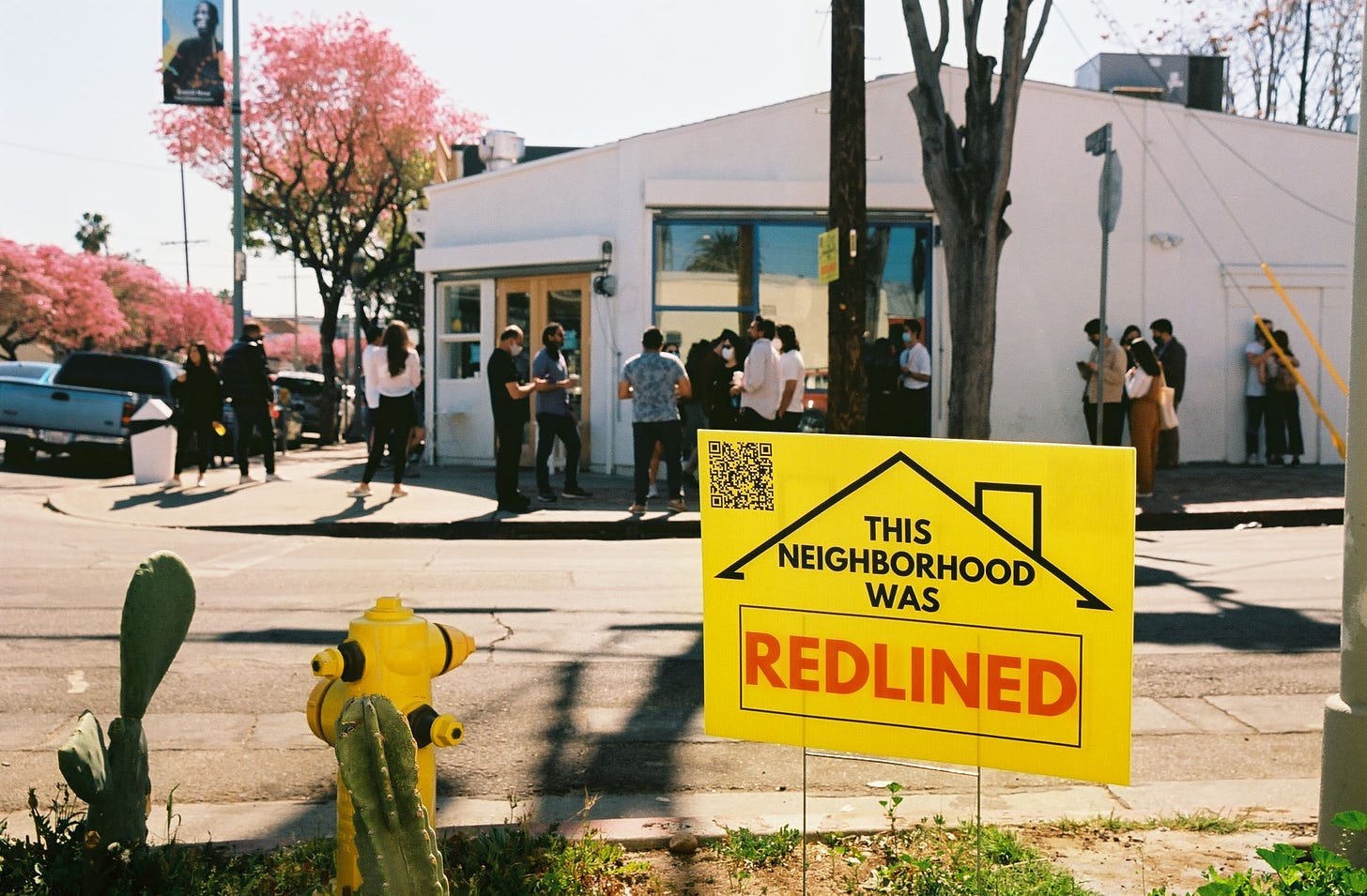 Photo of a yellow yard sign that says “This Neighborhood Was Redlined” planted in the dirt next to a cactus and a fire hydrant. Behind the sign is a long line of people waiting to order at Courage Bagel.
