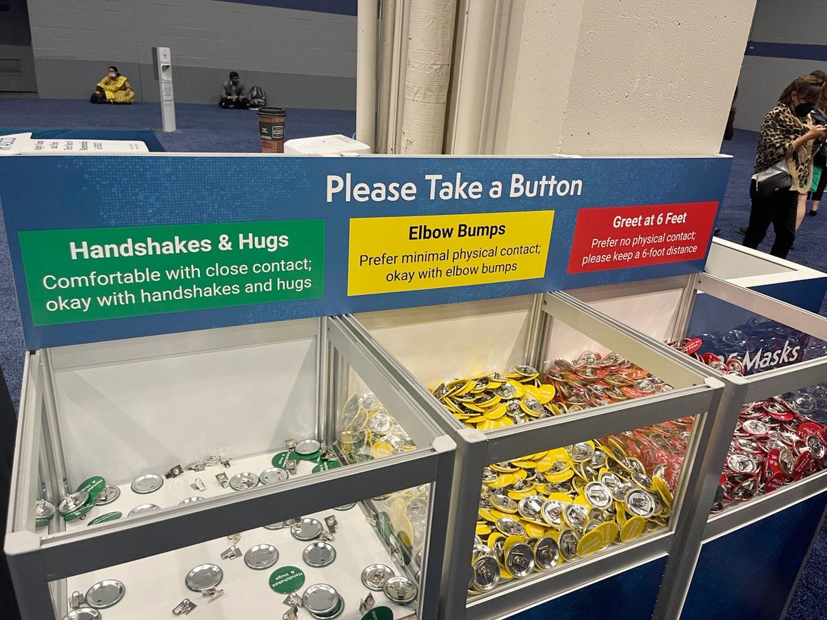 A sign says Please take a button, there are 3 bins, the first bin is labeled green and contains green buttons and the sign says Handshakes and hugs, another bin is full of yellow buttons and the yellow sign says elbow bumps, and the third bin is full of red buttons and is labeled great at 6 feet. In this picture the green bin is almost empty and the yellow and red bins are still quite full with buttons that nobody has taken