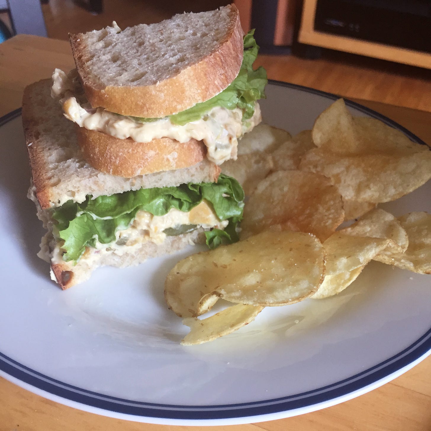 A plate with two sandwich halves stacked on top of each other, with green leaf lettuce and the chickpea filling falling out in places. A small pile of potato chips is next to them.
