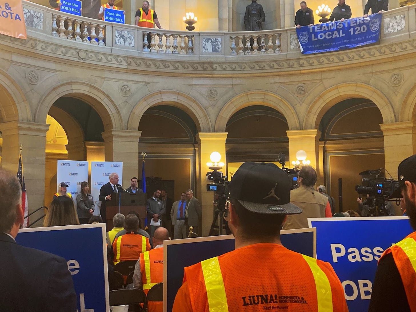 in the state capitol rotunda, governor walz stands at the podium in a suit, addressing a crowd of people wearing neon orange construction vests